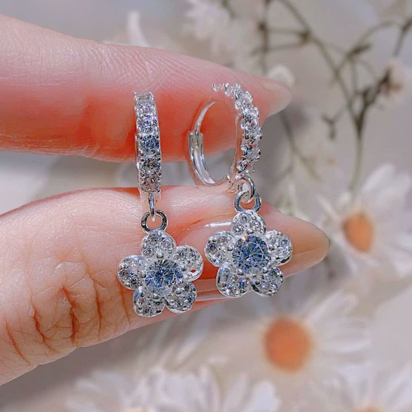 Flower-shaped silver earrings with clear stones held between fingers against a soft floral background.
