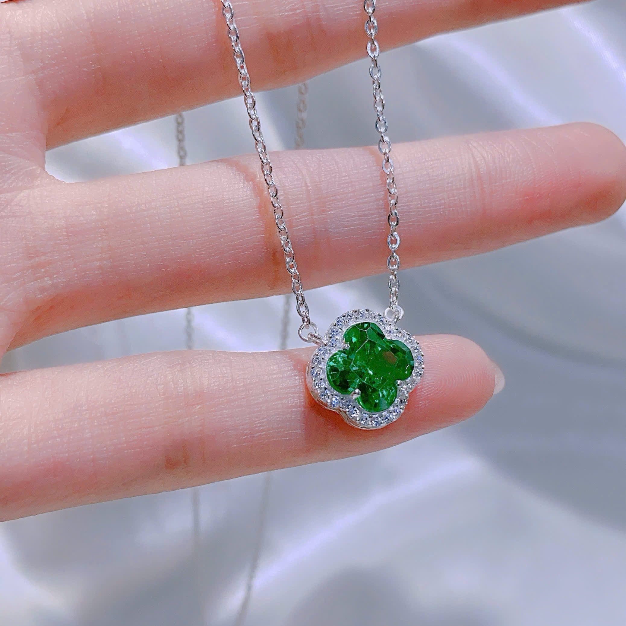 Green clover-shaped pendant on a silver chain held between fingers against a blurred background
