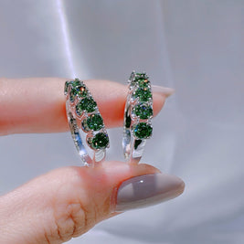 Green gemstone hoop earrings held between fingers against a neutral background