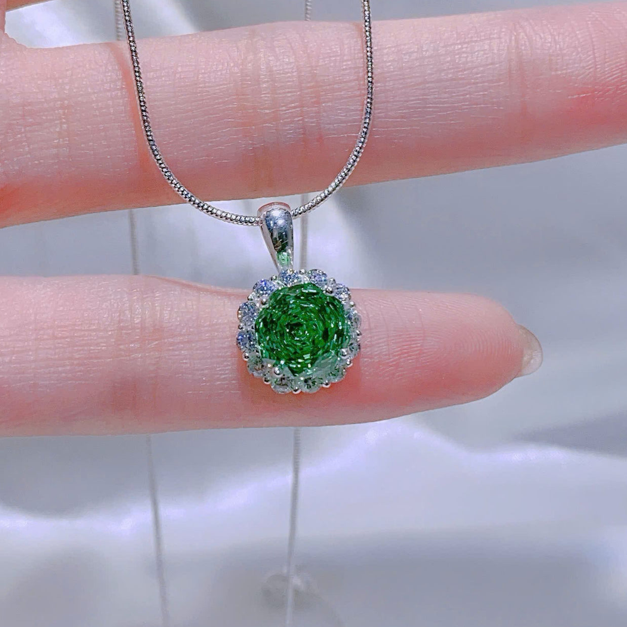Green gemstone pendant on a silver chain held by a hand against a white background