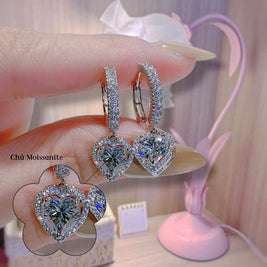 Heart-shaped diamond earrings held by a hand with a blurred background