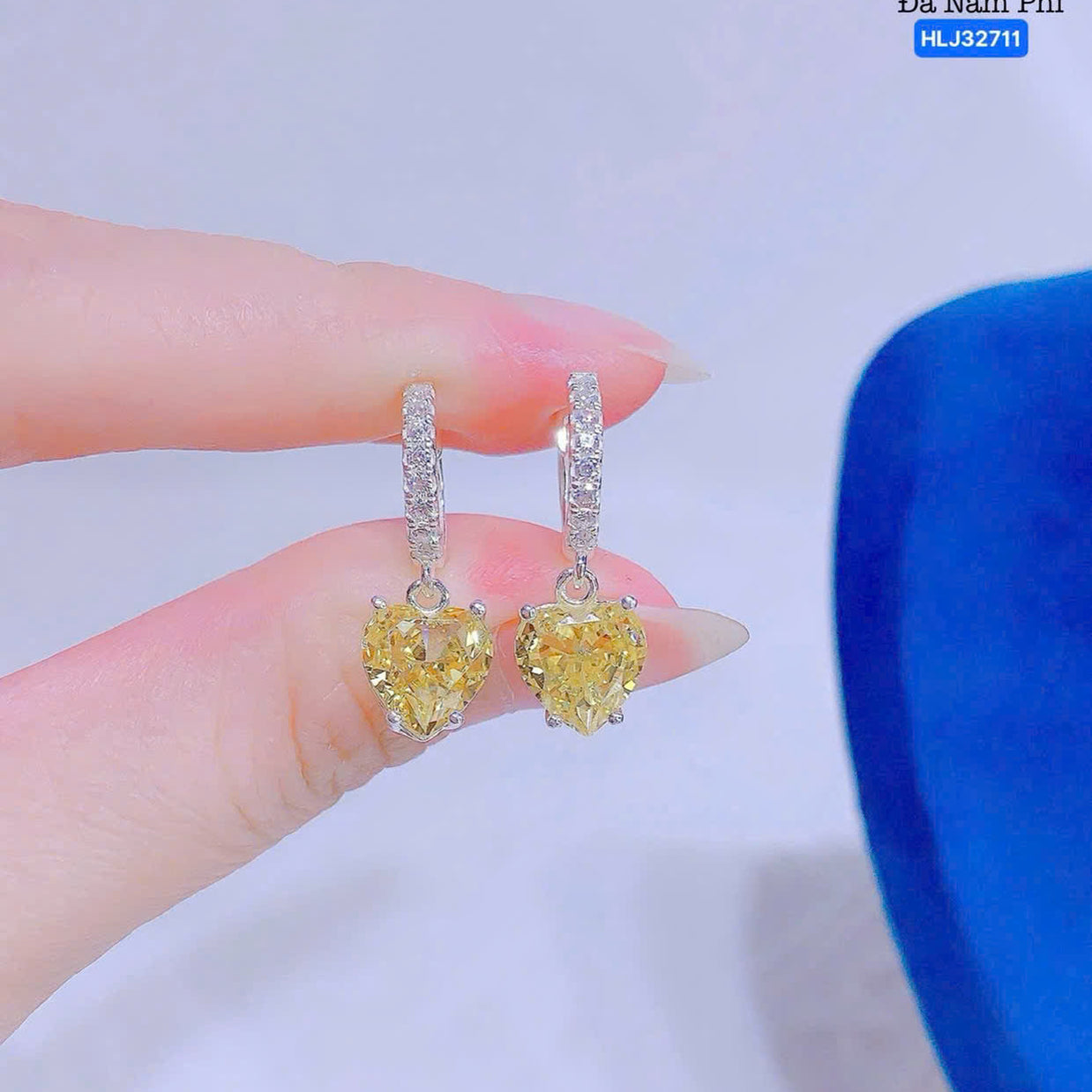 Heart-shaped yellow gemstone earrings held between fingers with a blue object in the background