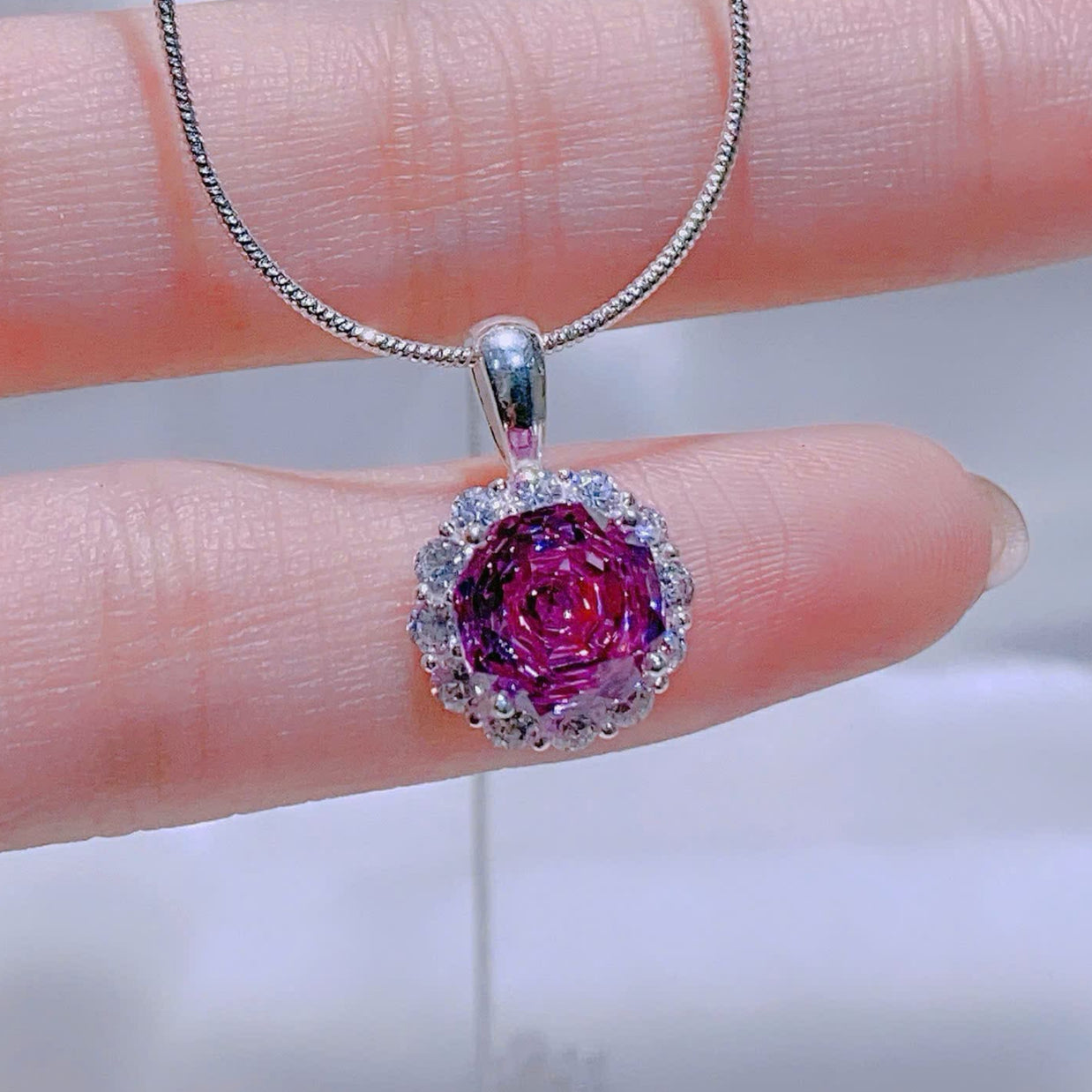 Pink gemstone pendant on a silver chain held by a hand against a blurred background