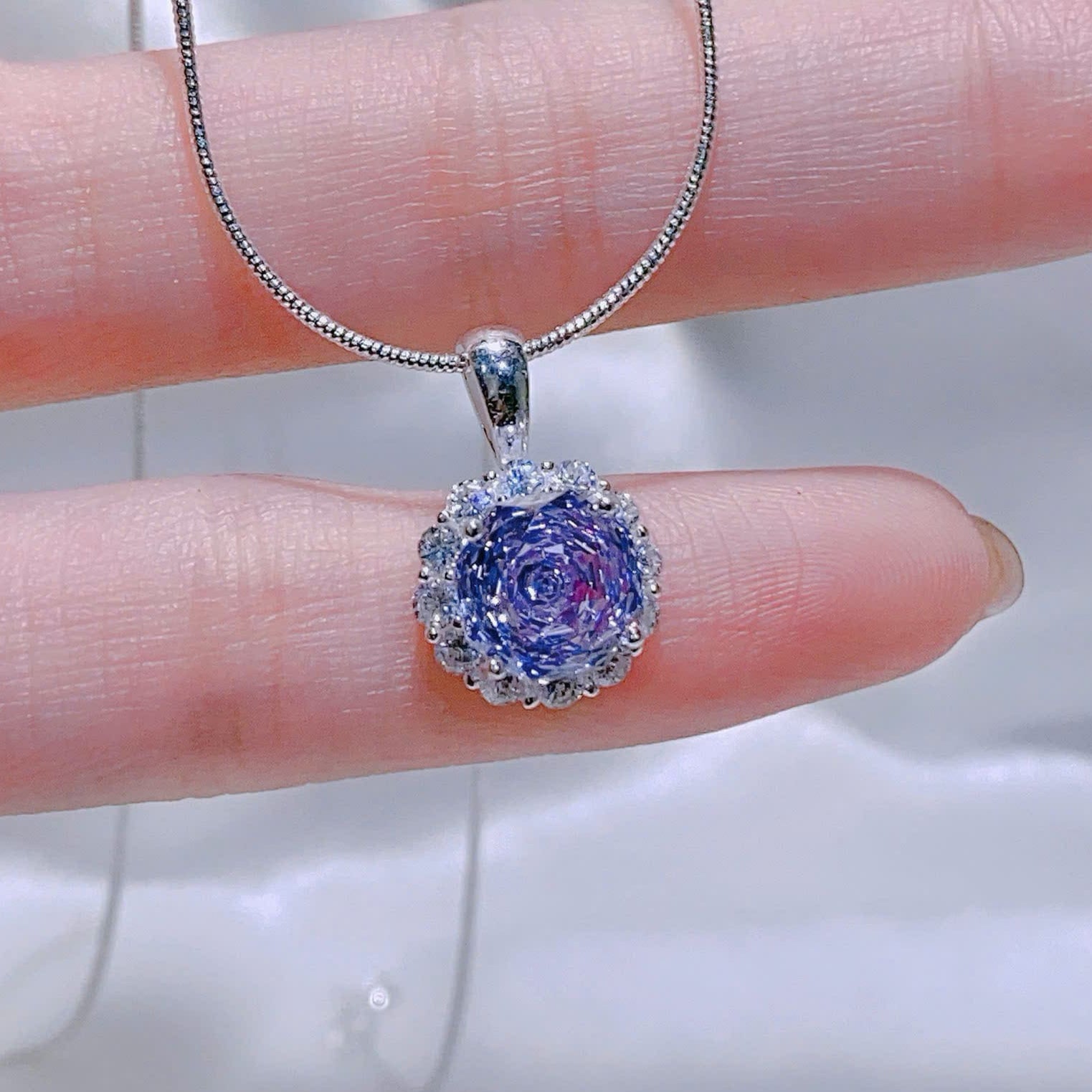 Purple gemstone pendant on a silver chain held by a hand against a white background