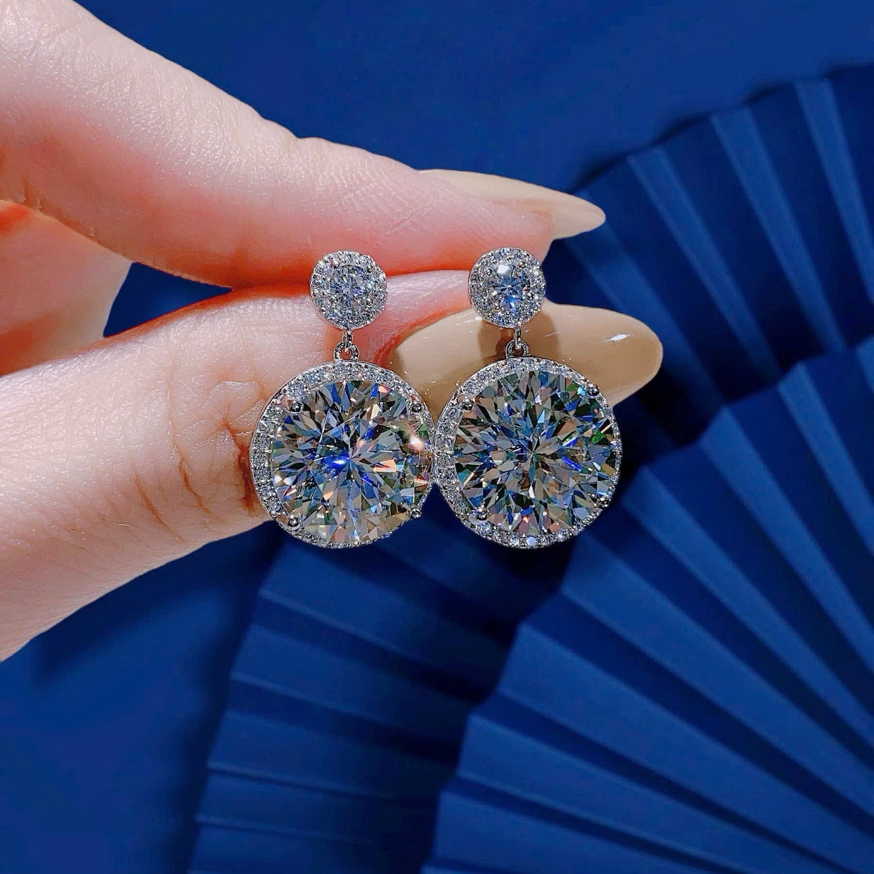 Round diamond earrings held by a hand against a blue background