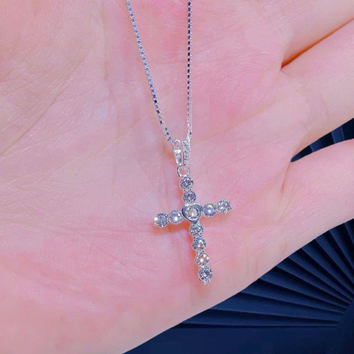 Silver cross pendant necklace held between fingers against a neutral background