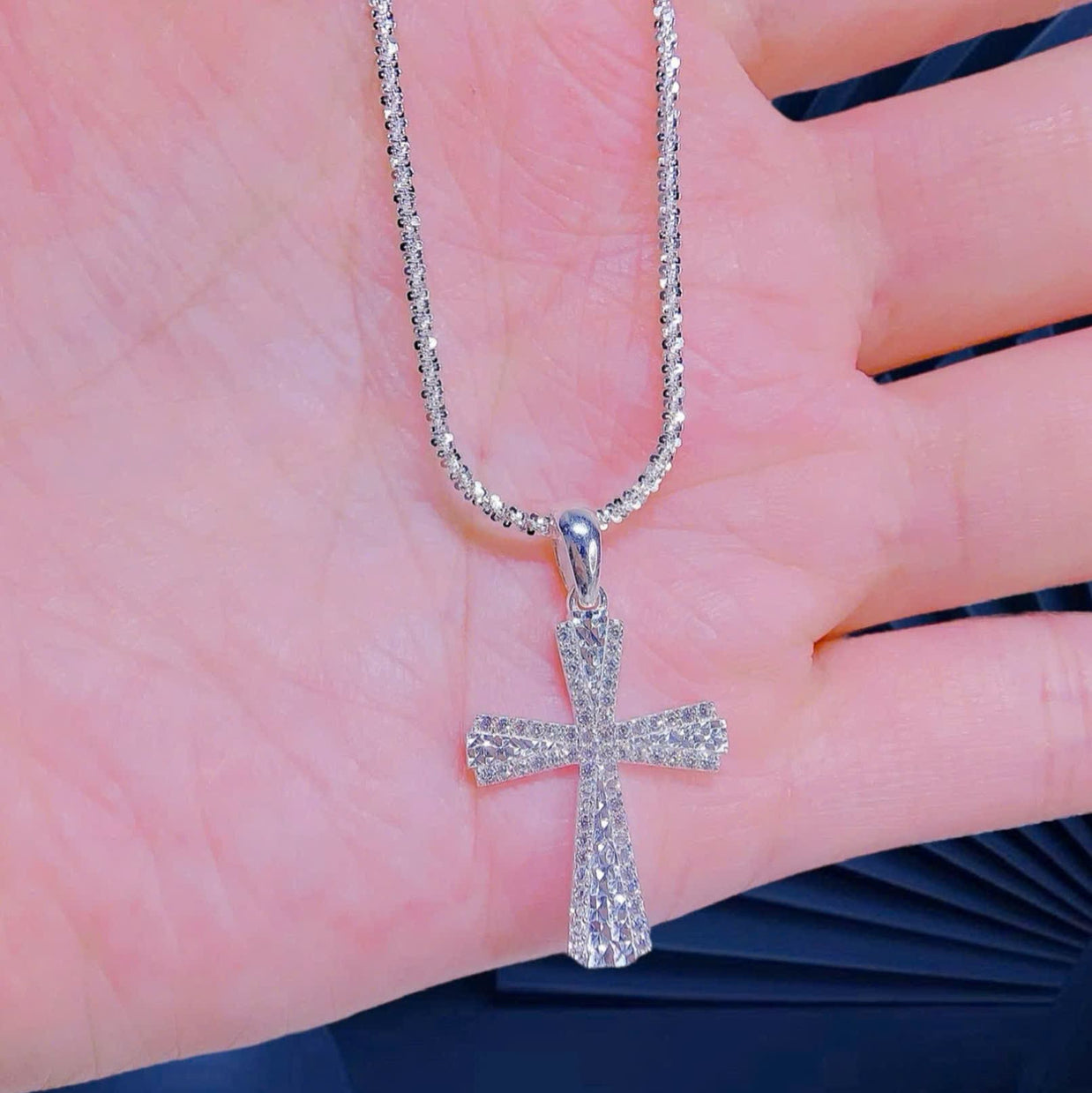 Silver cross pendant on a chain held against a person's hand.