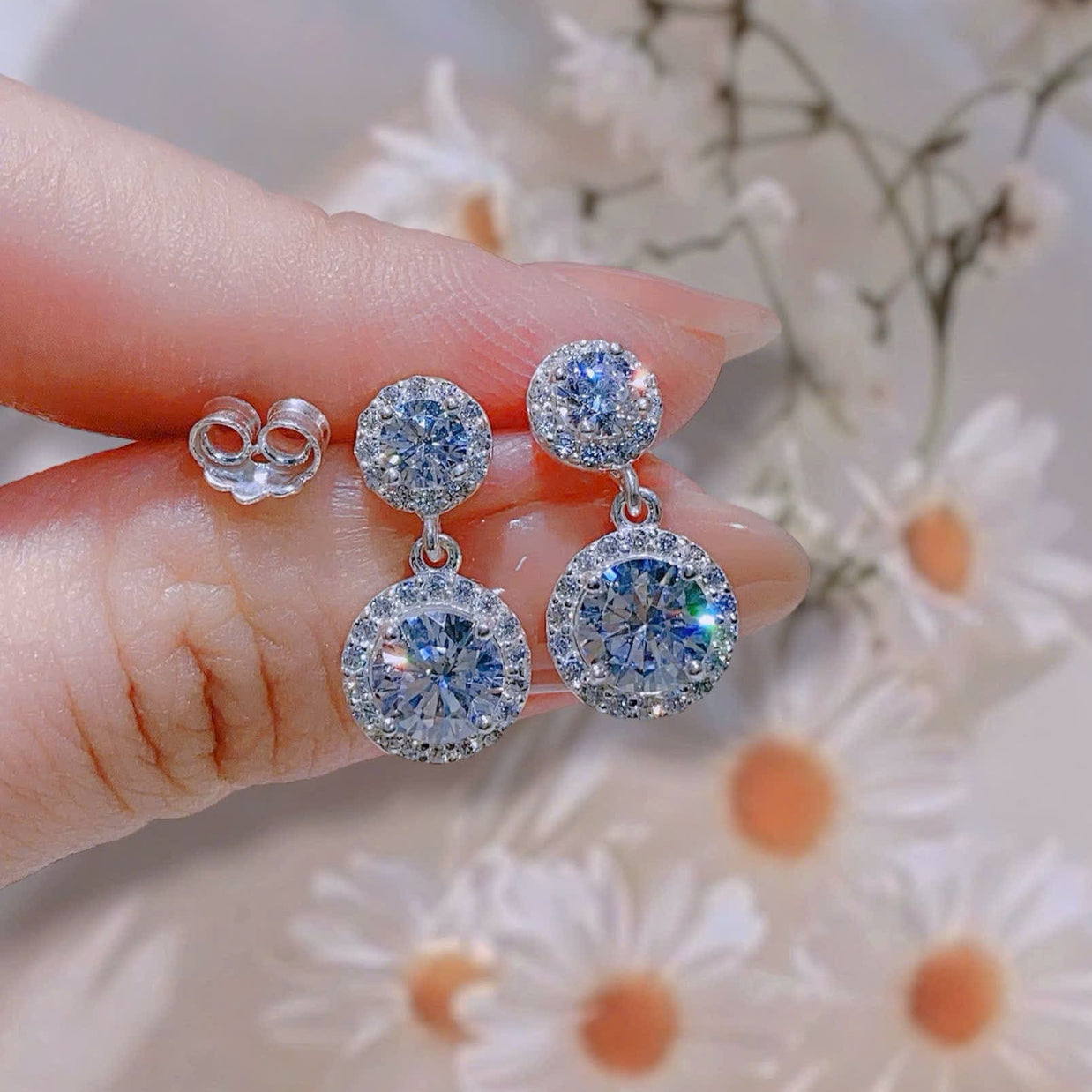 Silver earrings with clear stones held by a hand against a floral background