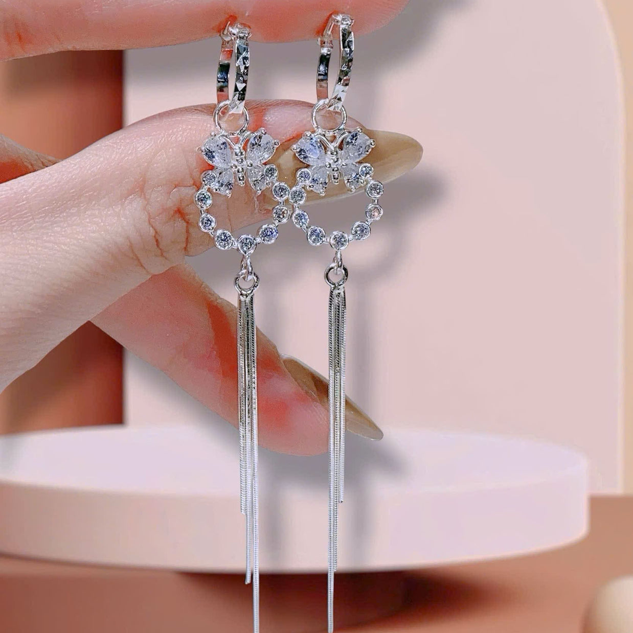 Silver earrings with heart-shaped designs and dangling elements held by a hand against a neutral background.