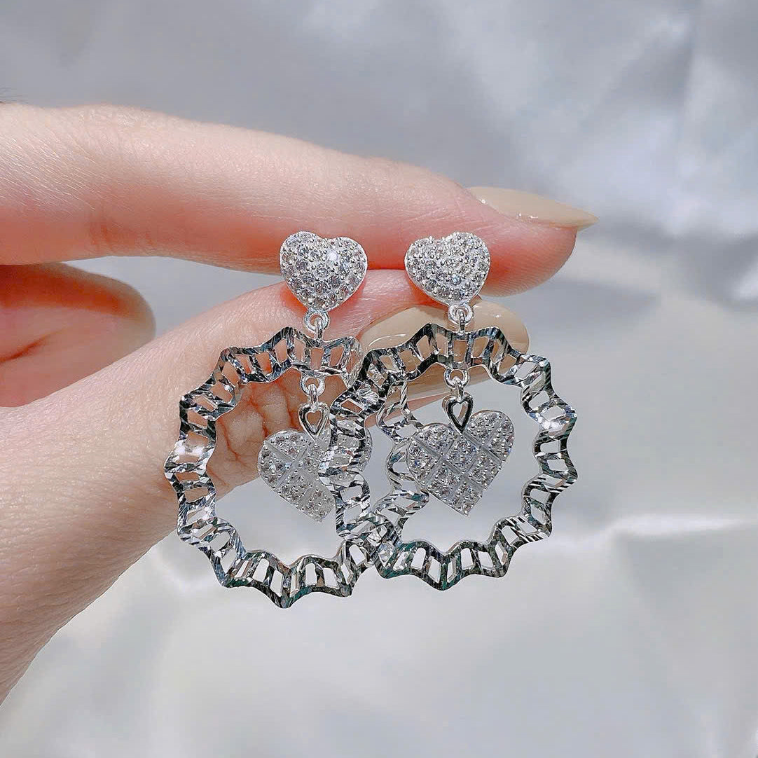 Silver heart-shaped earrings held by a hand against a neutral background