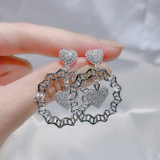 Silver heart-shaped earrings held by a hand against a neutral background