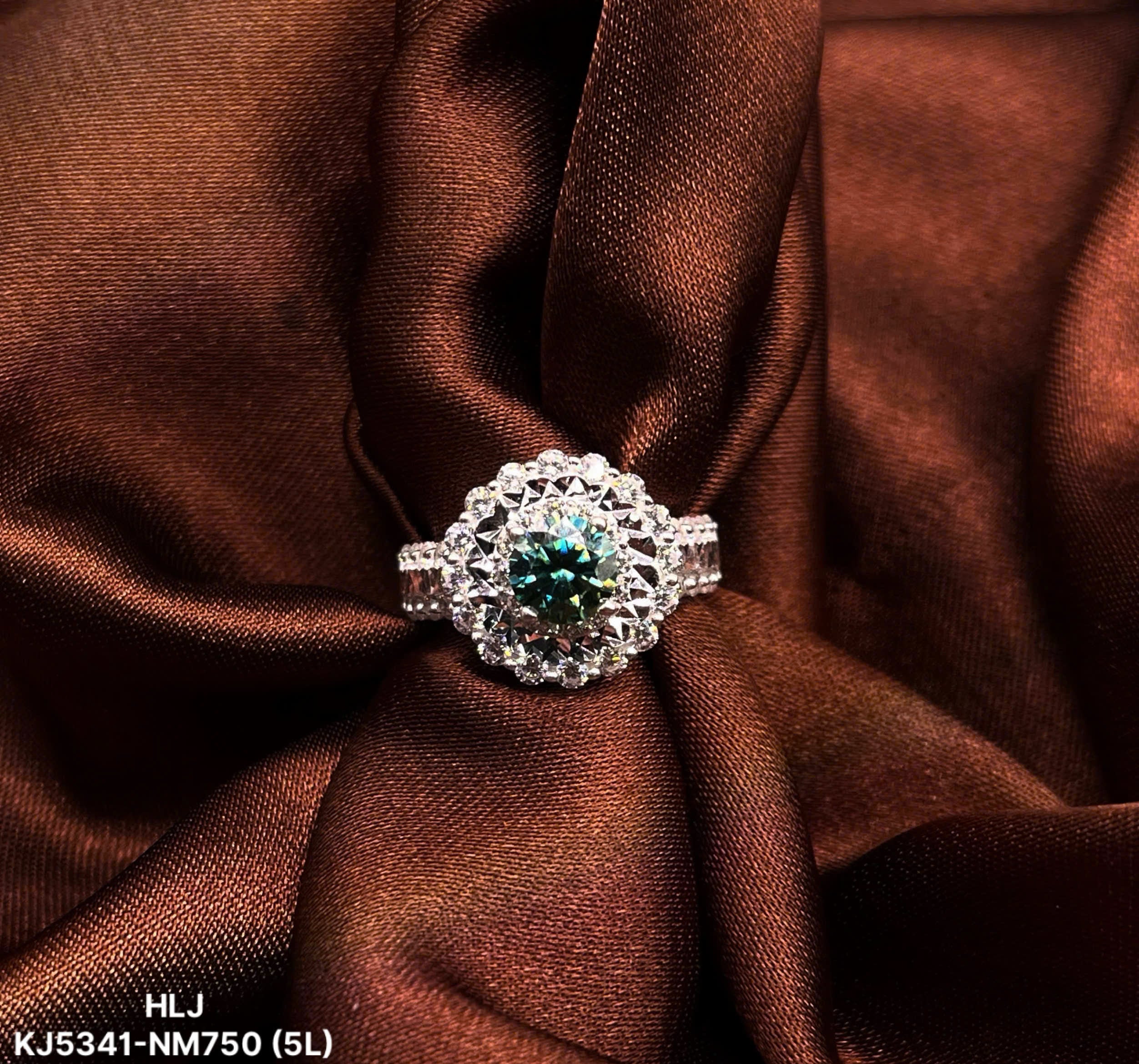 Silver ring with a green gemstone on brown fabric, brand 'HLJ' visible