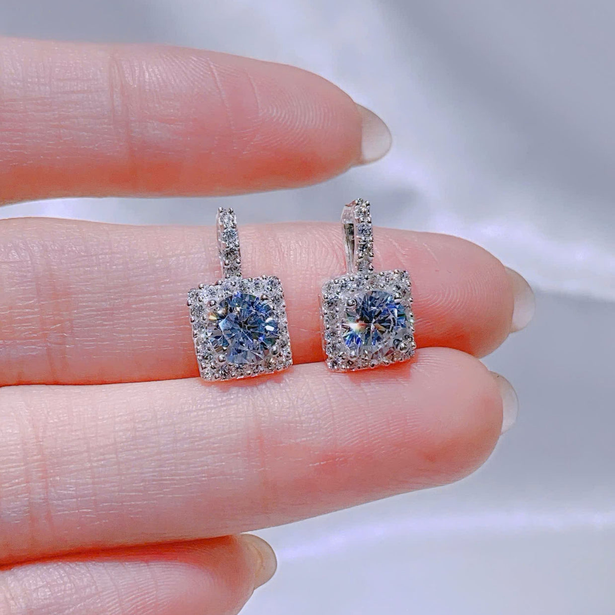 Square-shaped earrings with blue gemstones held between fingers against a neutral background